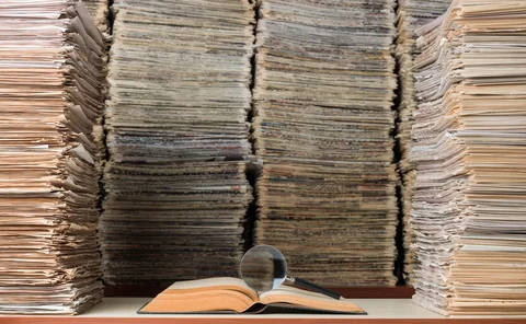 View of large amount of documents, newspapers, and books in classic library. A magnifying glass seen on top of an open book on desk. No people are seen in frame.