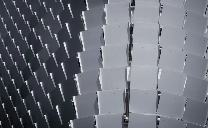 Abstract architectural pattern of repeating rectangular panels in alternating light and dark grey tones, creating a geometric 3D effect.