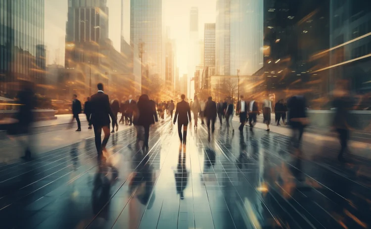 Blurred motion view of business professionals walking through a modern city street at sunrise, surrounded by tall glass skyscrapers reflecting golden light