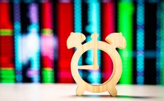 Wooden alarm clock icon placed on a table with colorful blurred stock market ticker screens in the background, representing time-sensitive trading, financial markets, and investment timing
