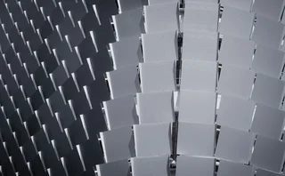 Abstract architectural pattern of repeating rectangular panels in alternating light and dark grey tones, creating a geometric 3D effect.