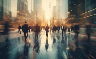Blurred motion view of business professionals walking through a modern city street at sunrise, surrounded by tall glass skyscrapers reflecting golden light