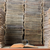 View of large amount of documents, newspapers, and books in classic library. A magnifying glass seen on top of an open book on desk. No people are seen in frame.